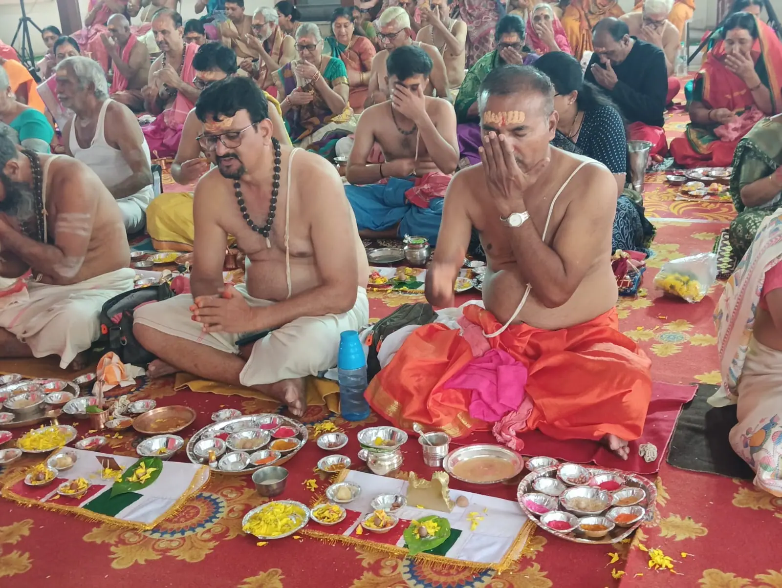 Men in saffron dhotis seated in rows, hands folded, with plates of offerings in front of them during Matru Shraddha